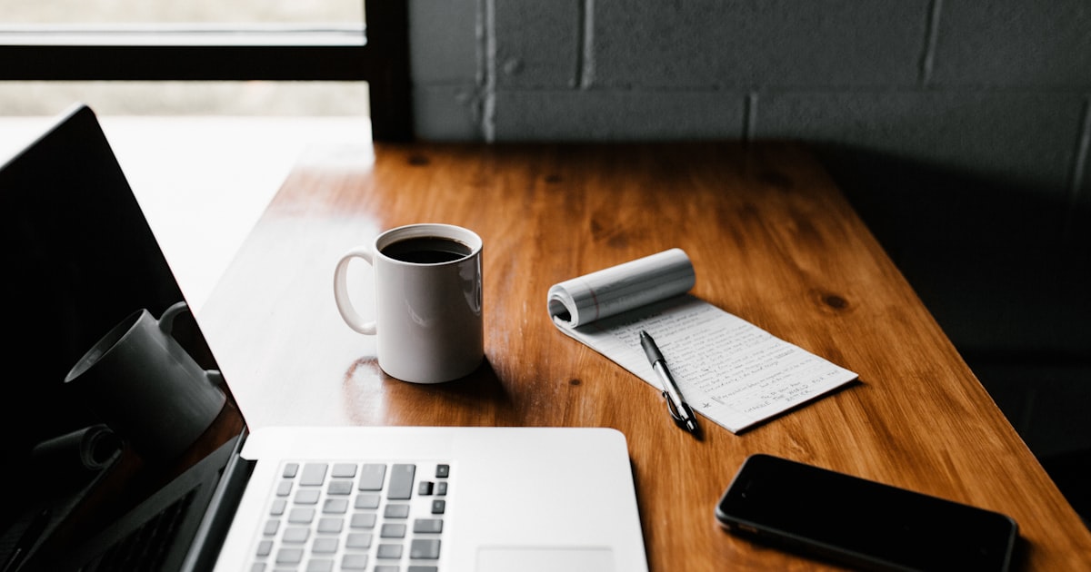 Warm beleuchteter Schreibtisch mit aufgeklapptem Laptop, Notizbuch mit Stift, Kaffeetasse und Smartphone – Sinnbild für Content-Recherche und GEO/SEO-Arbeit im Mittelstand